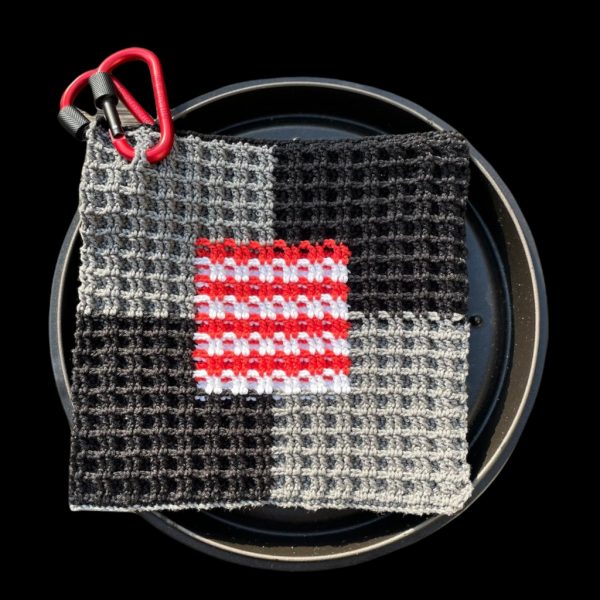 Handgemachter Topflappen im Patchwork-Design mit grauen und schwarzen Feldern sowie einem roten und weißen Muster in der Mitte, mit Karabinerhaken zum Aufhängen.