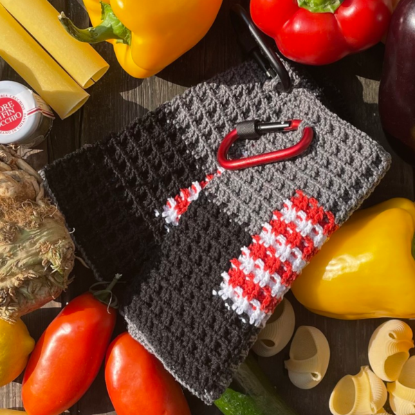 Handgemachter Patchwork-Topflappen mit grauen, schwarzen und einem rot-weiß karierten Feld, gehäkelt im Waffelmuster, mit rotem Karabiner zum Aufhängen.
