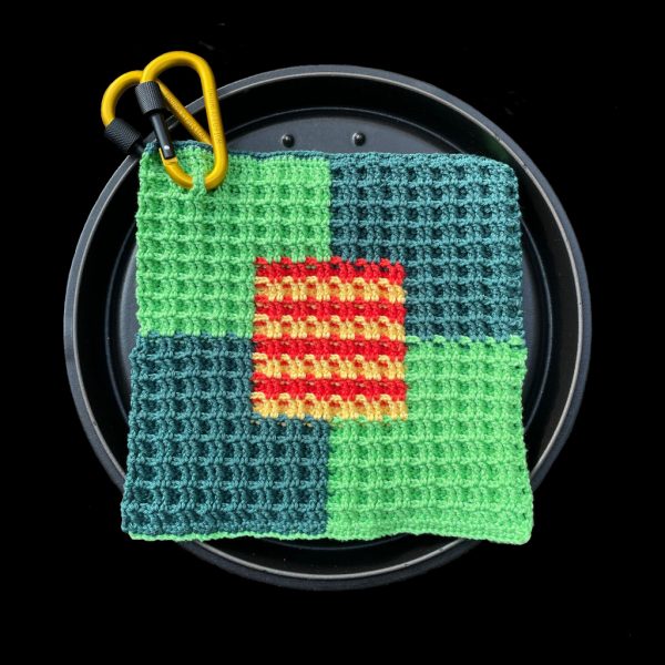 Quadratischer Topflappen mit dreidimensionalen Effekt. Zentriert in einem vier quadratischen Muster eingebettet befindet sich ein mit je vier Streifen abwechselnd rot. weiß gestreiftes Quadrat. Die vier umrandenden Quadrate sind farblich von oben links angefangen ein helleres Grün. Das Gleiche auch diagonal. oben rechts hat das Quadrat eine blau, graue Farbgebung, diagonal auch das Gleiche.